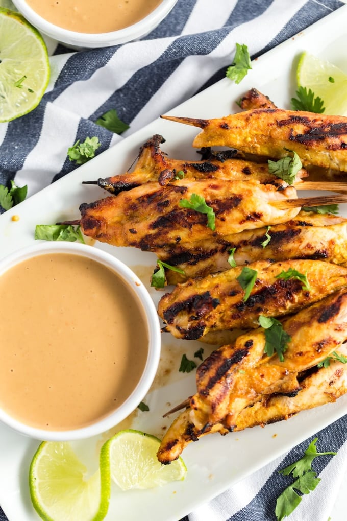 grilled chicken satay on a white platter with peanut sauce and a wedge of lime