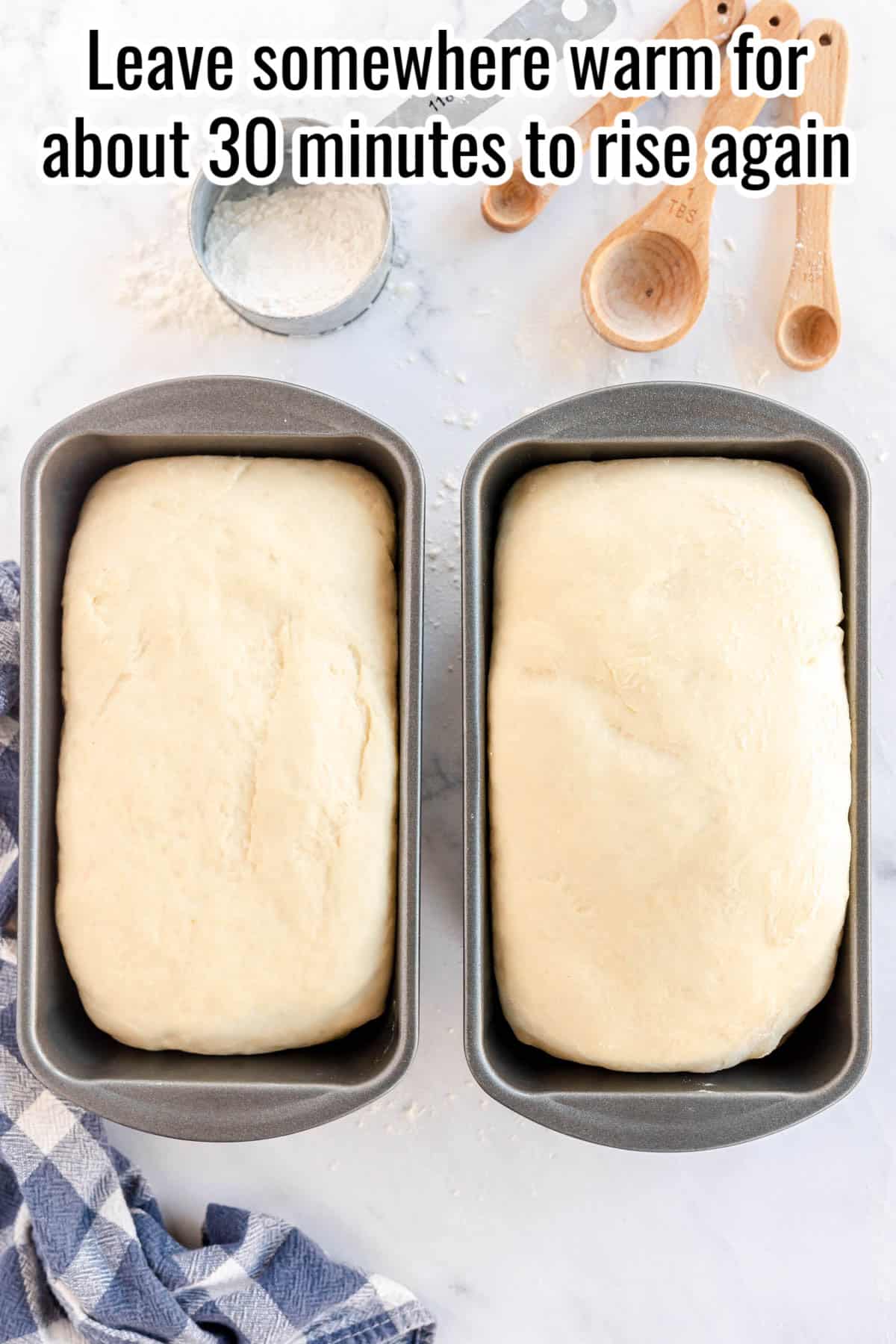 Two bread dough loaves in pans with a towel and utensils on a countertop. A caption above reads "Leave somewhere warm for about 30 minutes to rise again".
