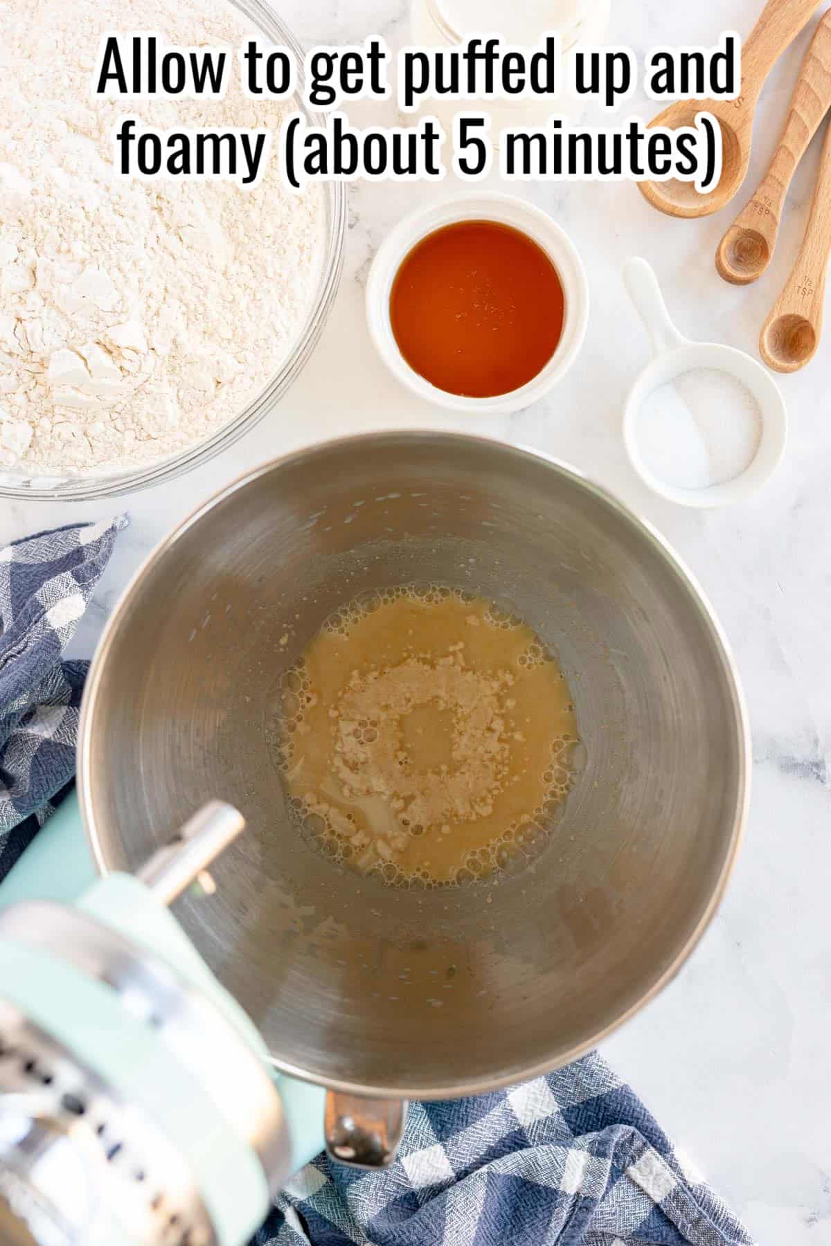 A mixing bowl with yeast mixture sits on a counter surrounded by flour, honey, salt, measuring spoons, and a blue checkered cloth. Text overlay reads, "Allow to get puffed up and foamy (about 5 minutes) for perfect sandwich bread.