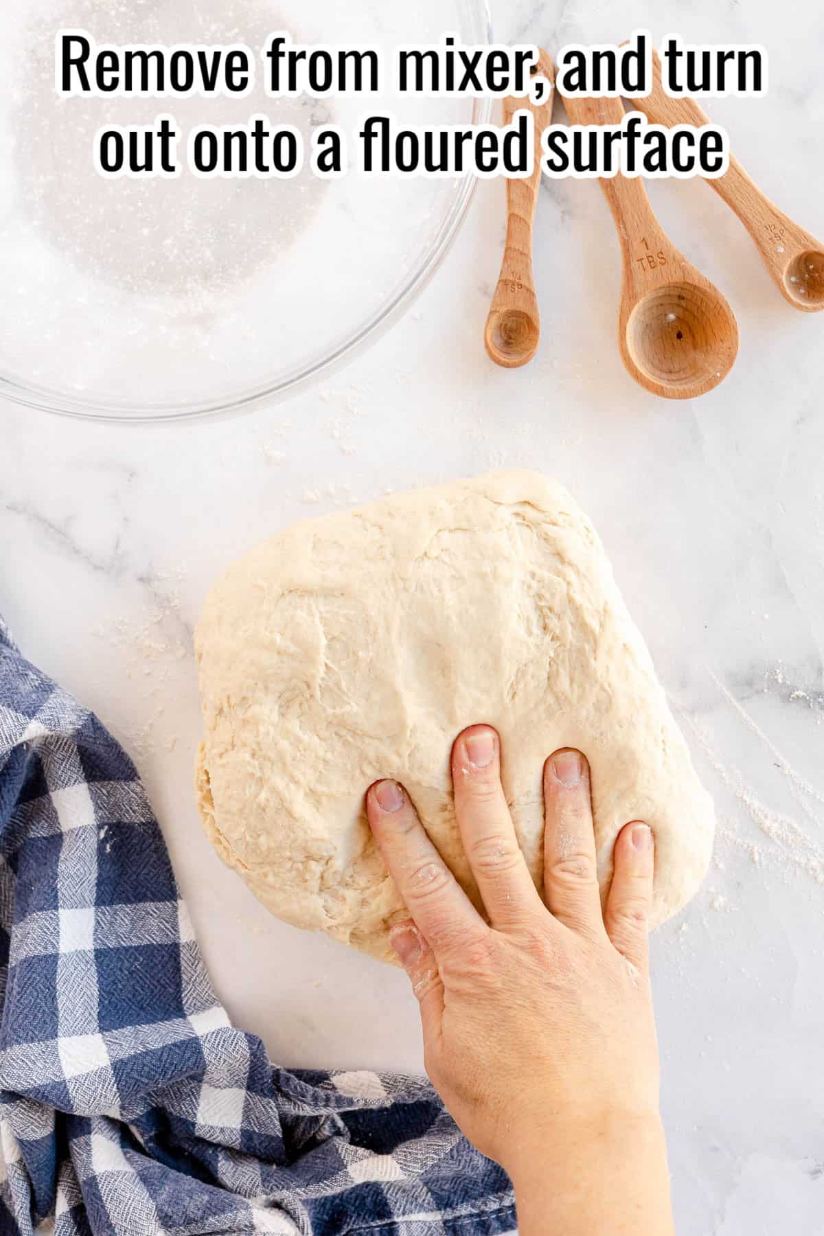 A hand is placing dough for sandwich bread onto a floured surface next to wooden measuring spoons and a checked cloth. Text at the top instructs to remove from the mixer and turn out onto the floured surface.