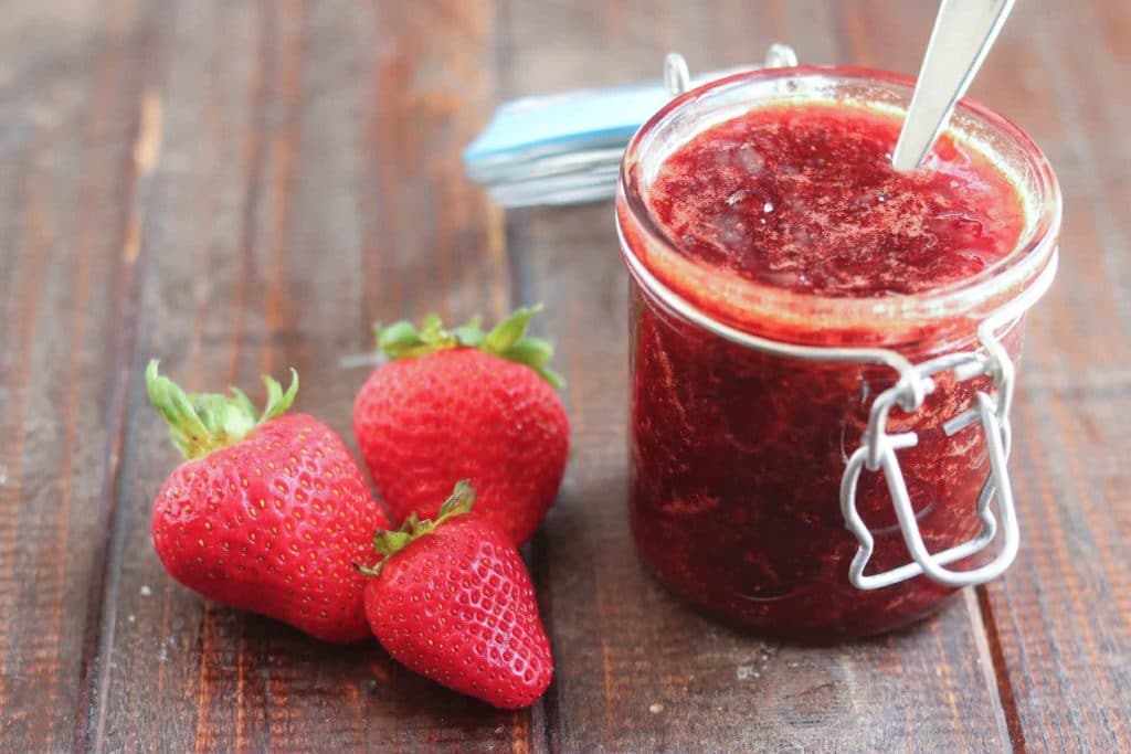 jar of strawberry jam with a spoon in it with strawberries alongside it